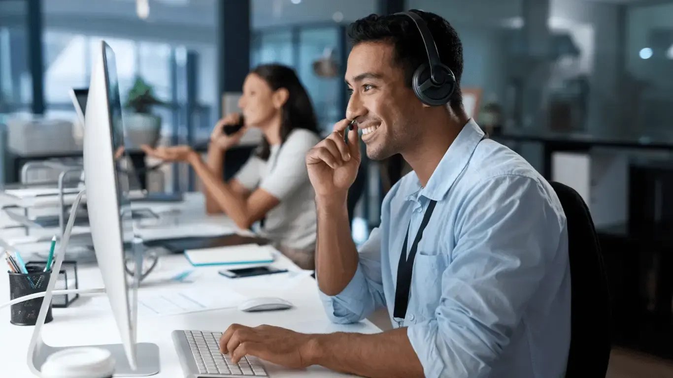 Man with headset looking at a desktop computer with a female colleague on the phone behind him out of focus.